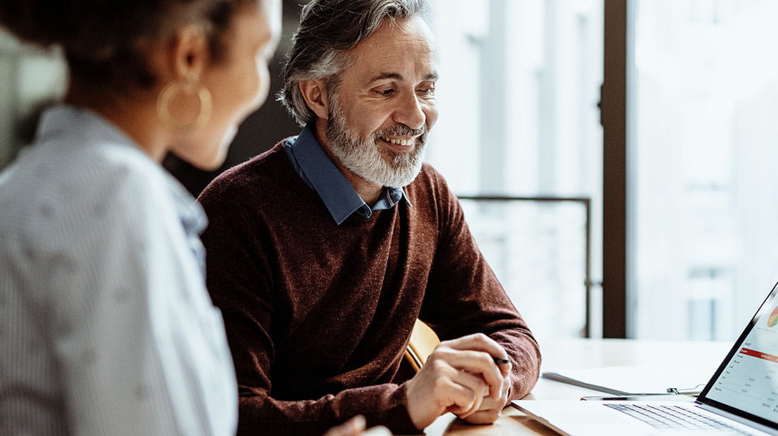 An older man with grey hair and a beard sits with a young woman with dark curly hair. They sit at a desk looking at data on a laptop.