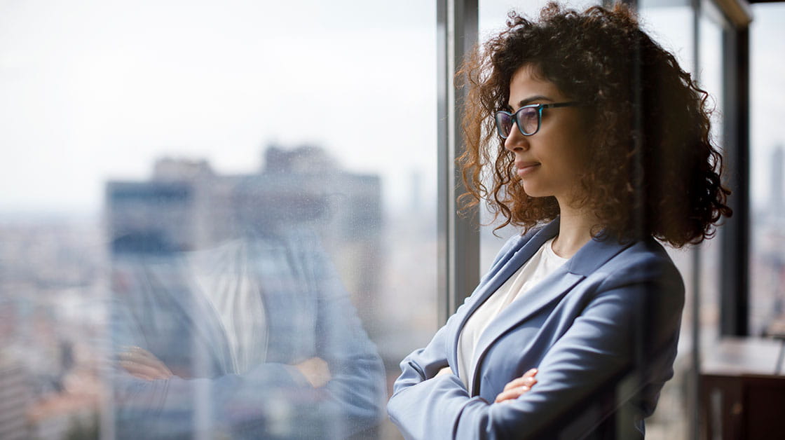 A professional woman stands looking out onto the skyline. She has long brown curly hair and wears glasses, a grey suit and a white shirt.