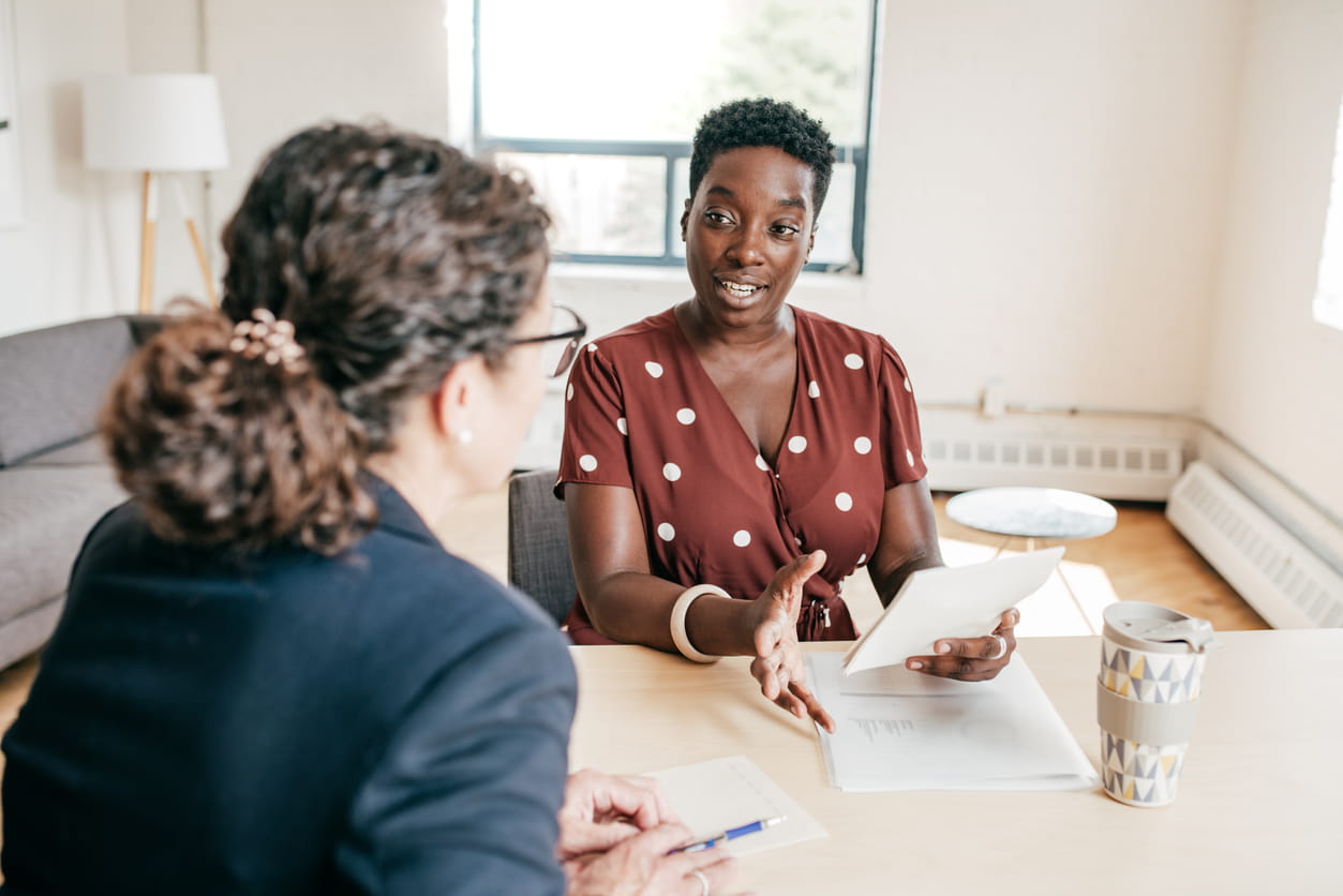 A Black woman is sitting at a table holding a notepad. She is wearing a brown dress with white polkadots. She is talking to a white woman wearing a black jacket, also sitting down at the table.