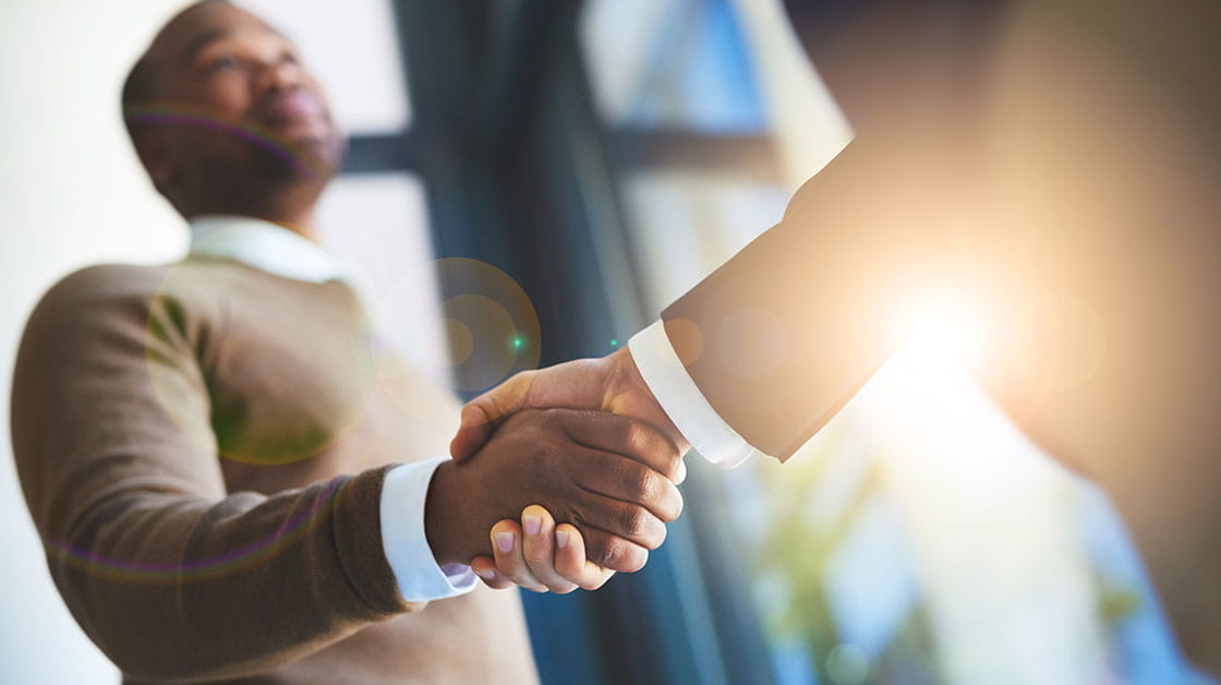 Two men shake hands: a black man with short black hair and a closely cropped beard wearing a beige jumper and white shirt, and a white man wearing a dark suit and white shirt.