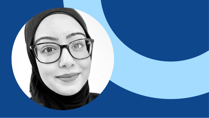 A black and white photo on a blue background of an Asian woman wearing a head scarf and glasses. She is smiling with her mouth closed. 