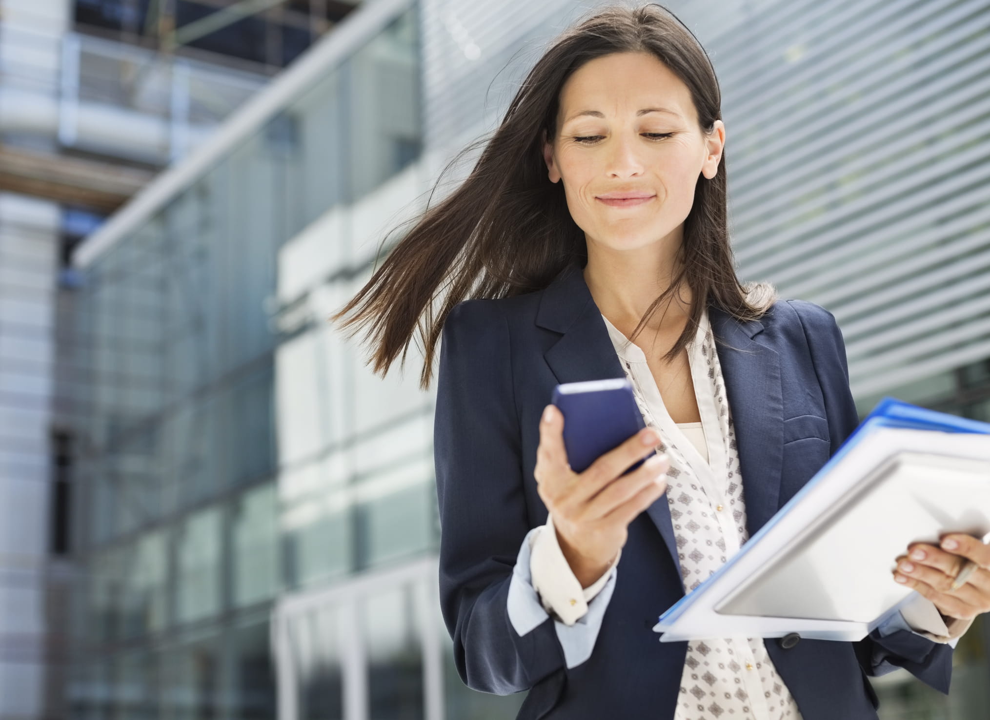 A white woman with long brown hair wearing a blue suit and white shirt looks at her phone. She is smiling with a closed mouth and holding a folder. 