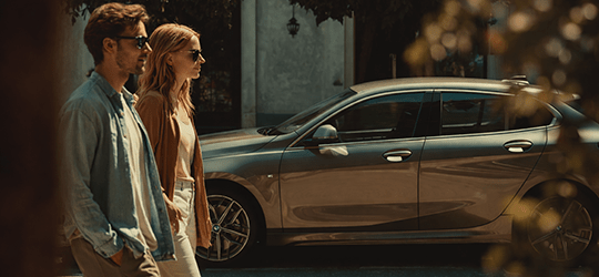 A man and woman in sunglasses walk past a gray parked car