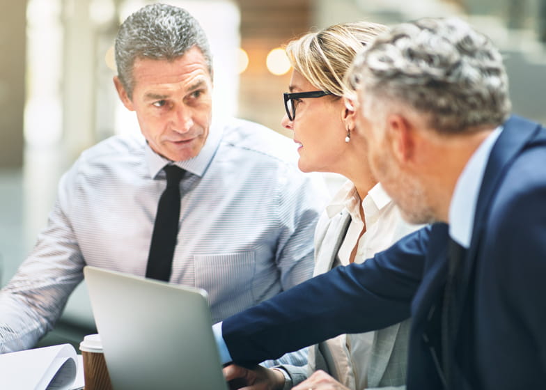 Three professionals are standing in front of a computer, talking. A white man with grey hair, wearing a blue striped shirt and navy blue tie is on the left. A white woman with blonde hair and glasses, wearing a grey jacket, white blouse and dangly earrings is standing in the middle. A white man with grey hair is standing on the right, wearing a blue suit, pointing to something across them.