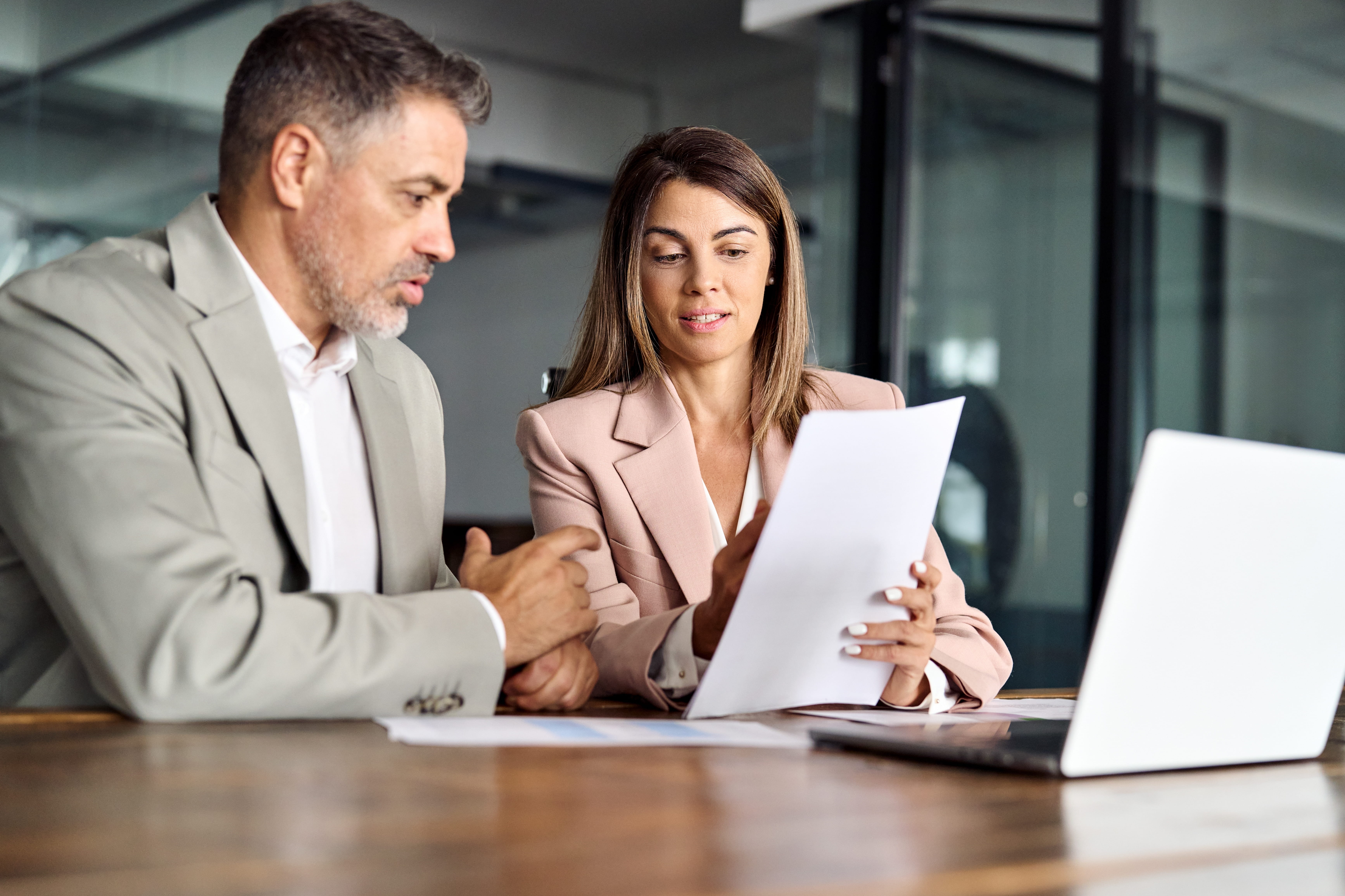 Two professionals are sitting at a desk looking over a paper. There is a laptop in front of them. A white man in a beige suit jacket is in the foreground and a white lady with brown hair is sitting next to him. 