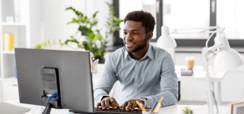 A black man with a bear, wearing a blue shirt is sitting at a desk typing on a desktop computer. There is also a white lamp on the desk. There is a big green plant behind and a white desk with a white lamp behind him. 