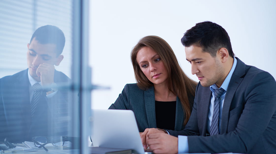 Two men and women professionals are sitting in an office looking at a computer. The man is wearing a navy blue suit and the woman is wearing a blue jacket and black top.
