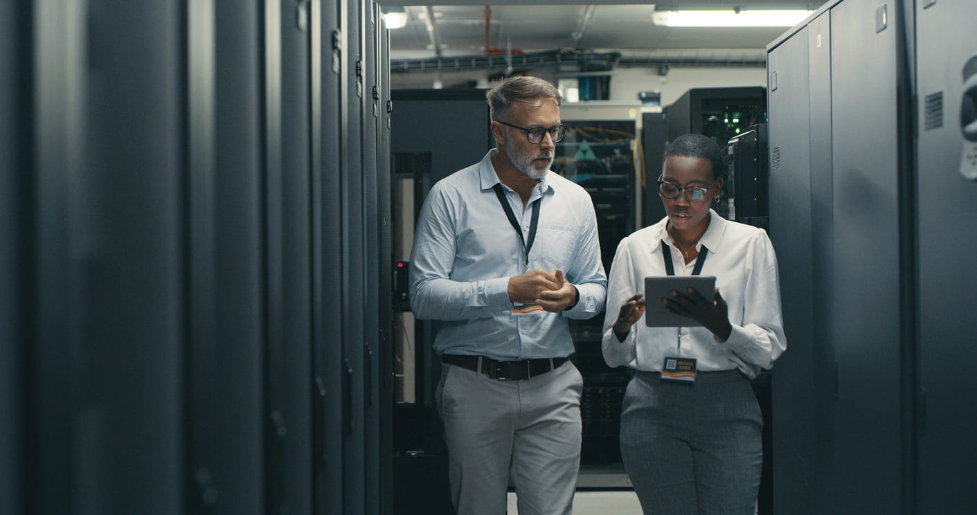 A black woman and a white man are walking through an office. They are both wearing white shirts and grey trousers. They have passes around their necks and the woman is holding an ipad. 