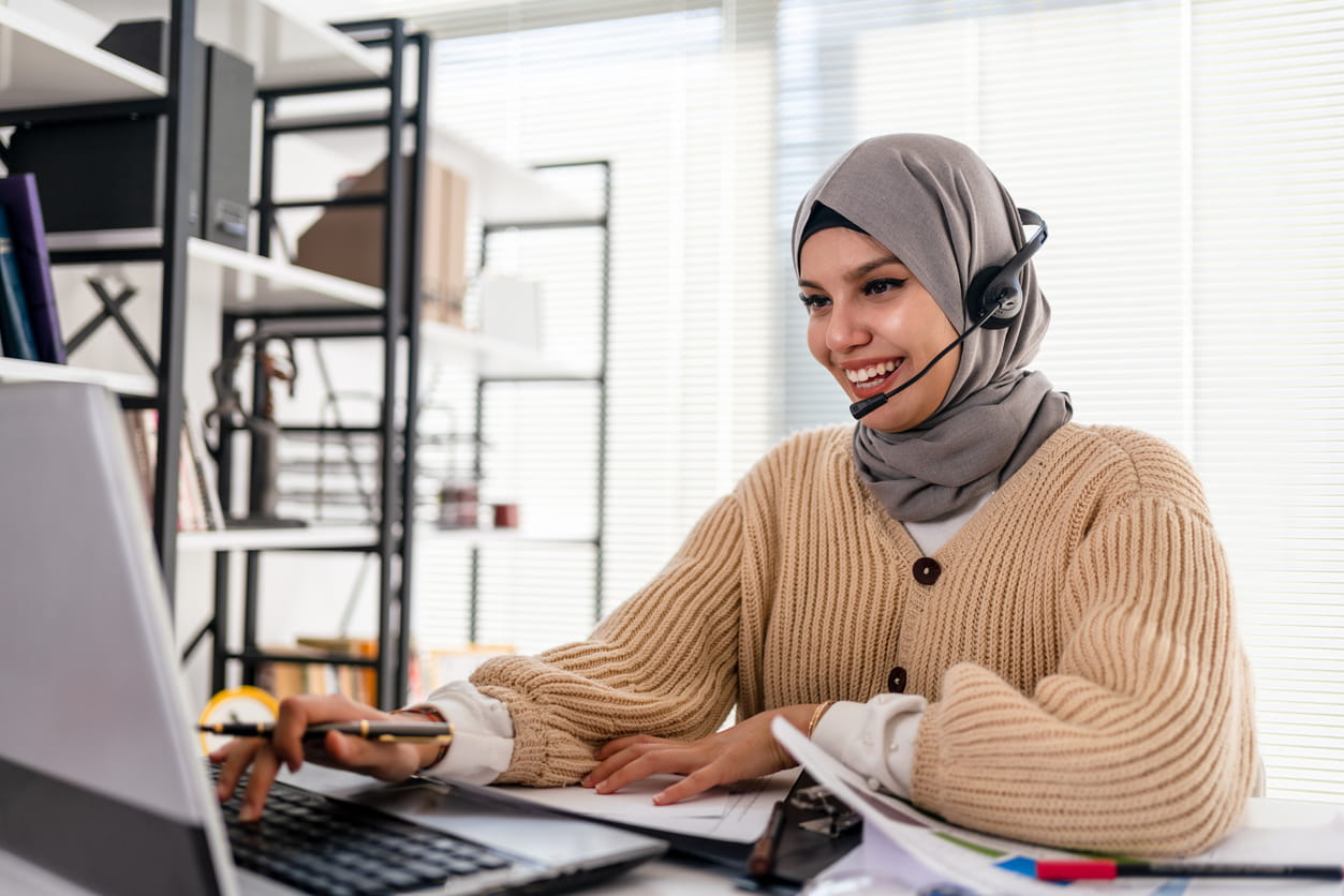 A woman wearing a hijab and beige cardigan is working at her computer wearing headphones.