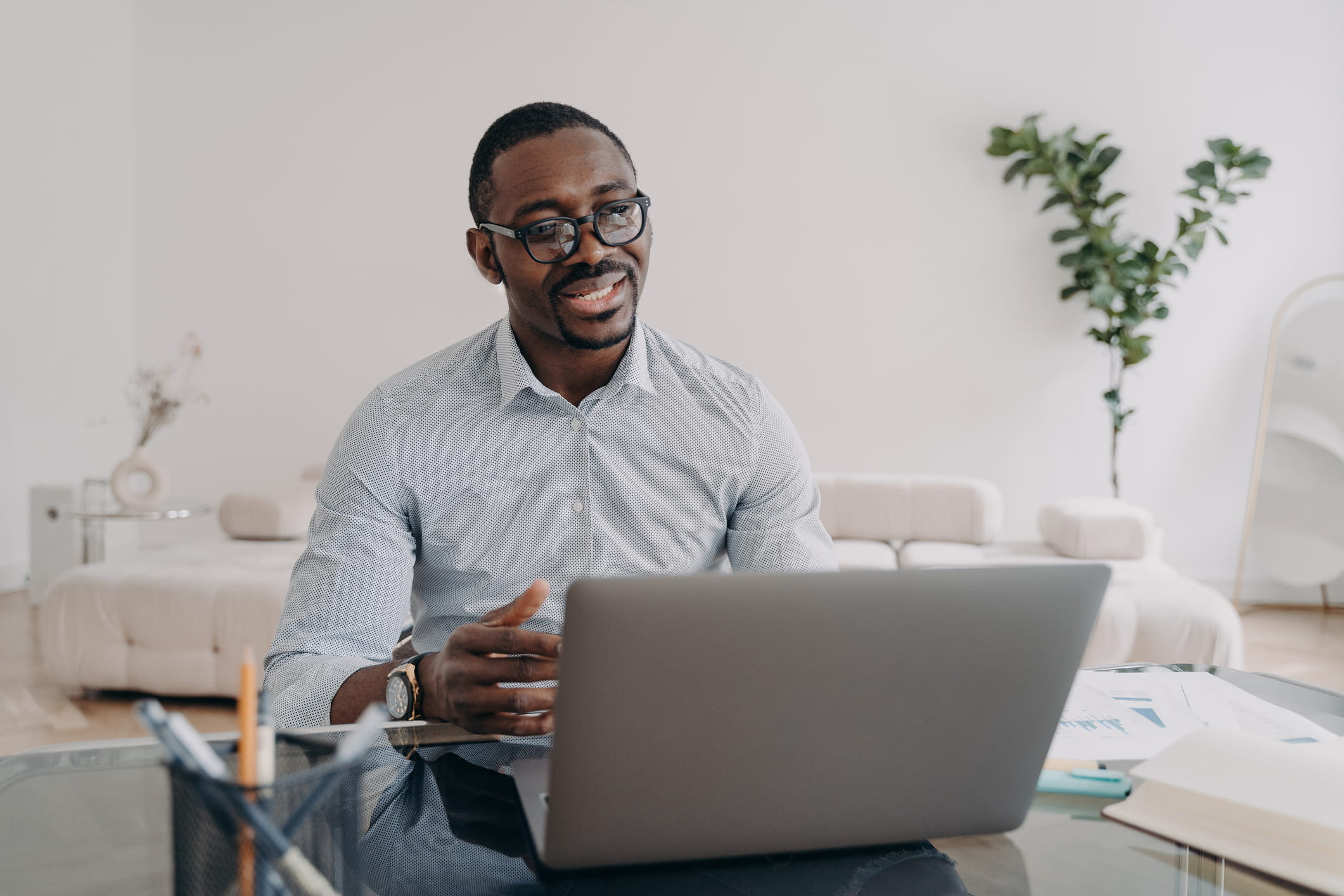 A black, male professional is sitting at a desk, looking at a laptop. He is wearing glasses and is explaining something to someone on the screen.