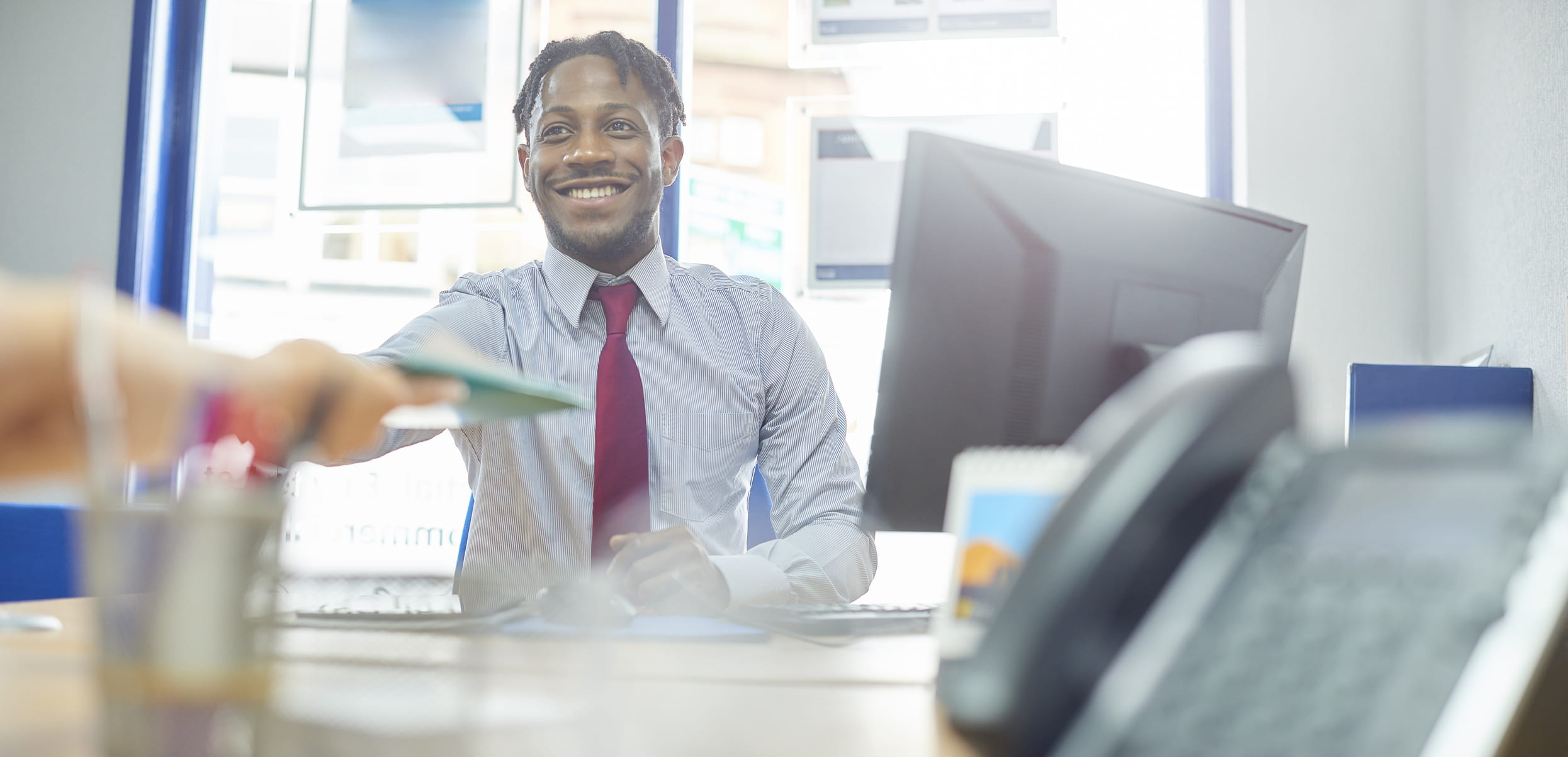 A young, black male professional is sitting at his desk. He is wearing a blue shirt and red tie and is smiling. He is taking a file off someone else. 