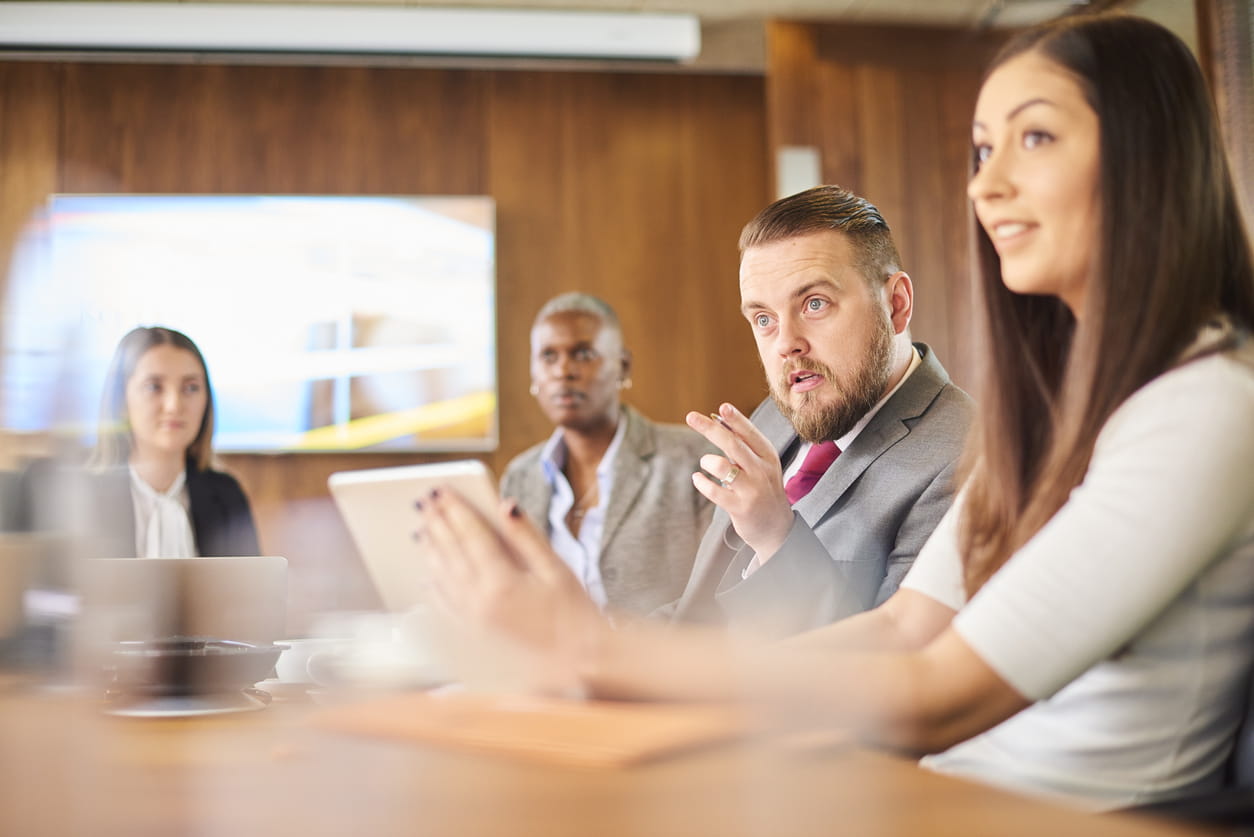Four professionals are sitting in a meeting room. A white man is talking, gesticulating and staring intently. He is sitting next to a white woman with long brown hair. 
