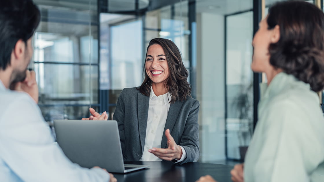 A white woman is sitting in an office at a desk, wearing a grey jacket and white shirt. She has a short brown bob and is smiling.