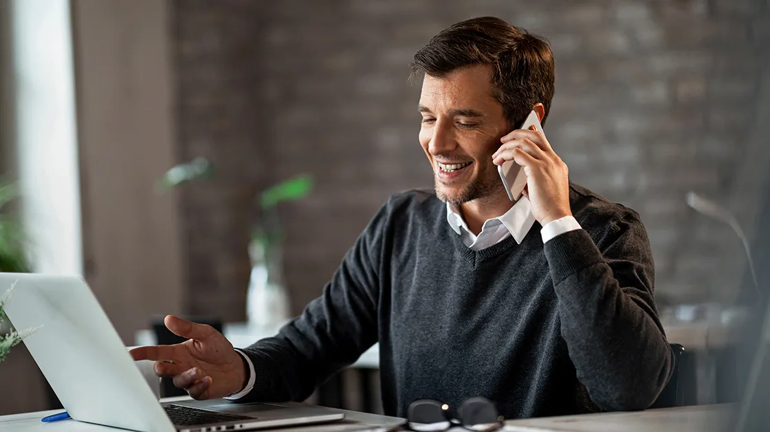 A white man with short, dark brown hair smiles while talking on a mobile phone. He sits in a modern office, next to an open laptop, which he gestures at as he talks.