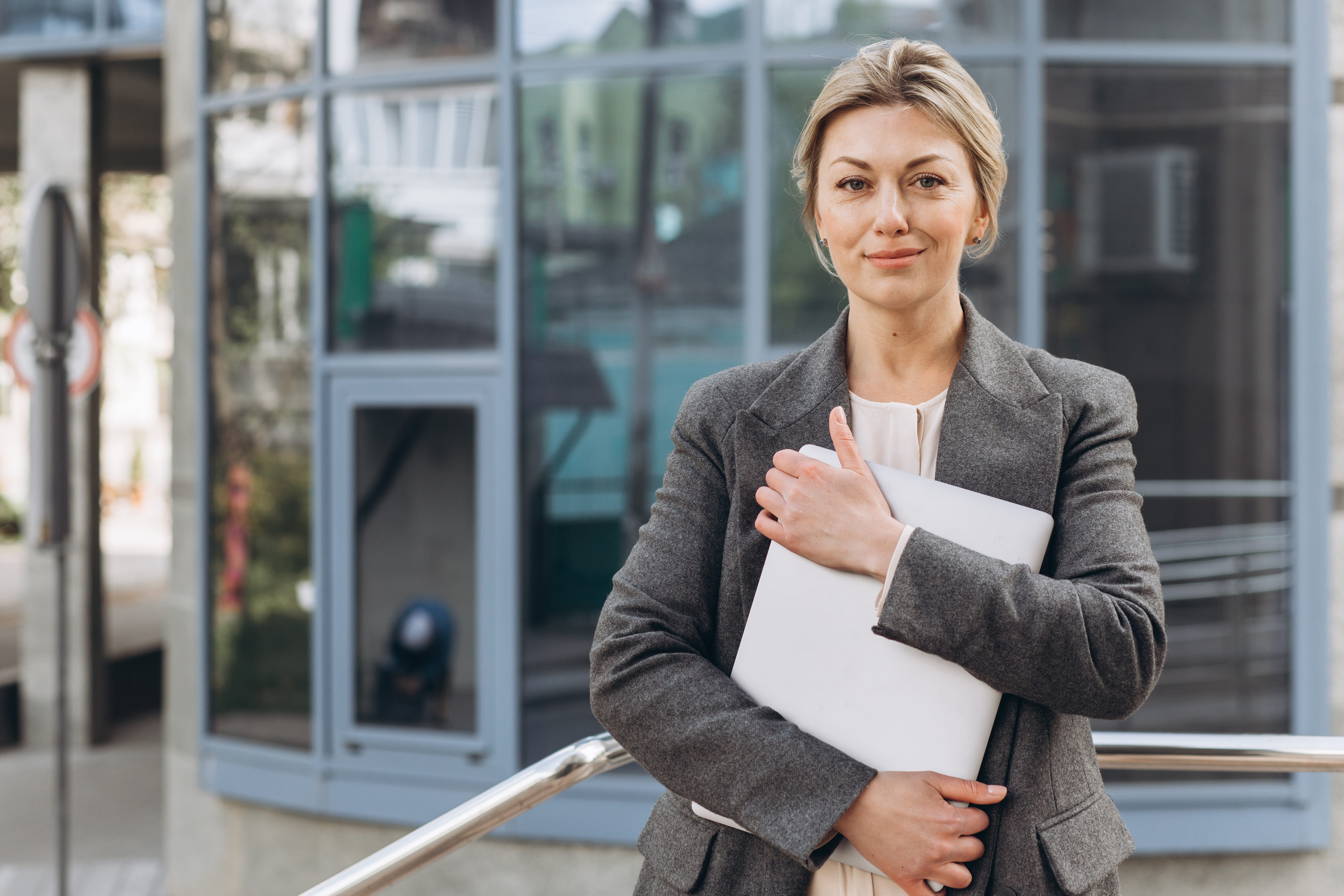 A white woman with blonde hair is standing outside an office building. She is wearing a grey jacket and holding papers in her arms. She is smiling with her mouth closed. 
