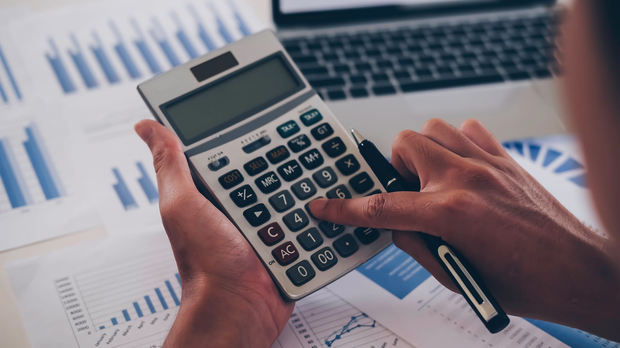 A person typing into a calculator, holding a pen. A laptop and papers on a desk.