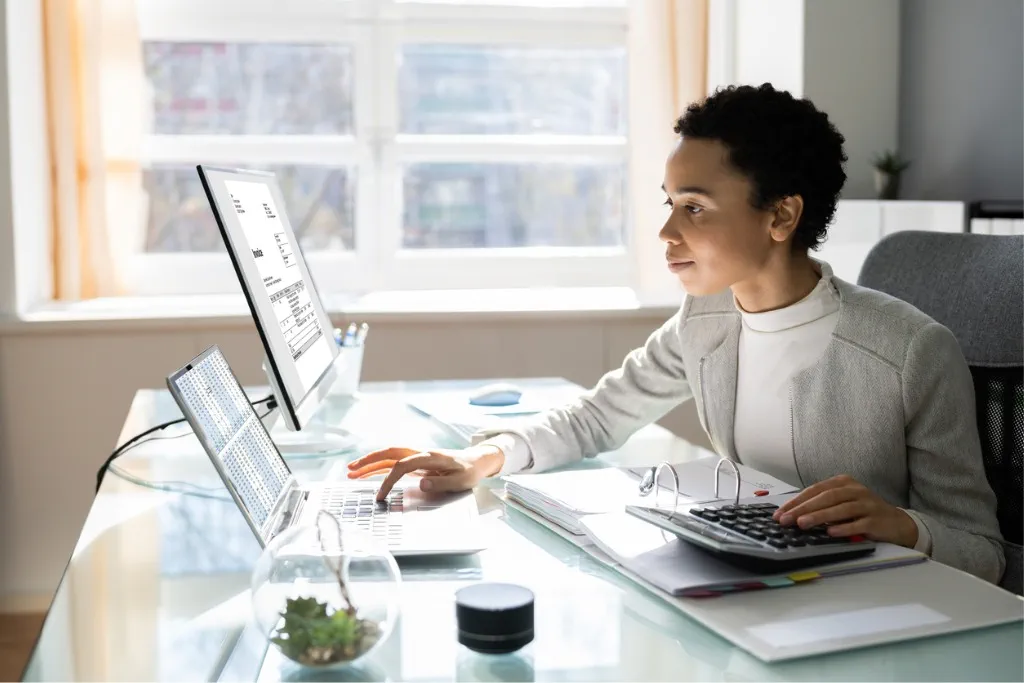 A black woman with short black hair is wearing a grey jacket and white top. She is working in front of two computer screens.