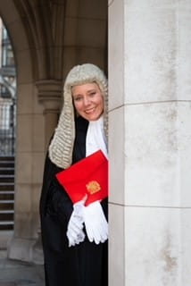 Amanda Brown QC is a white woman, wearing a Queen's Counsel's black robes and white wig. She's holding an embossed, red folder and looking out from behind a stone archway, smiling at the camera.