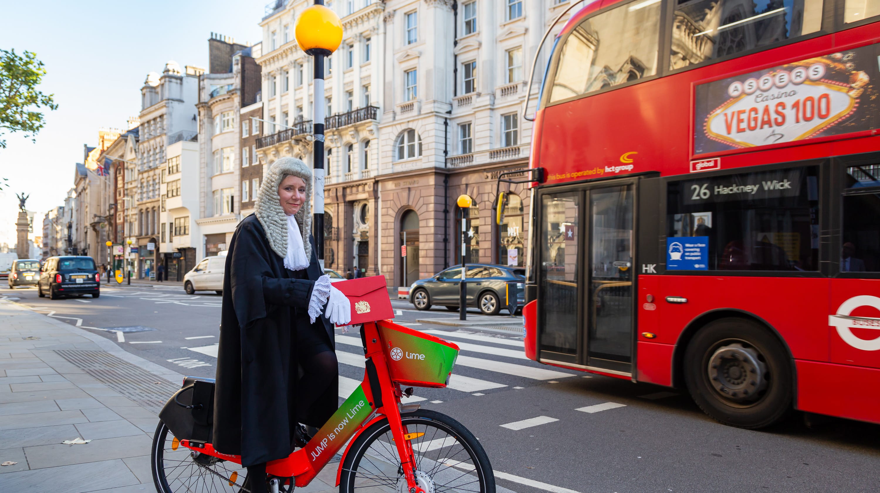Amanda Brown QC rides a red, electric hire bike on a London street. She is a white woman, wearing a Queen's Counsel's black robes and white wig. She's holding an embossed, red folder and smiling at the camera.