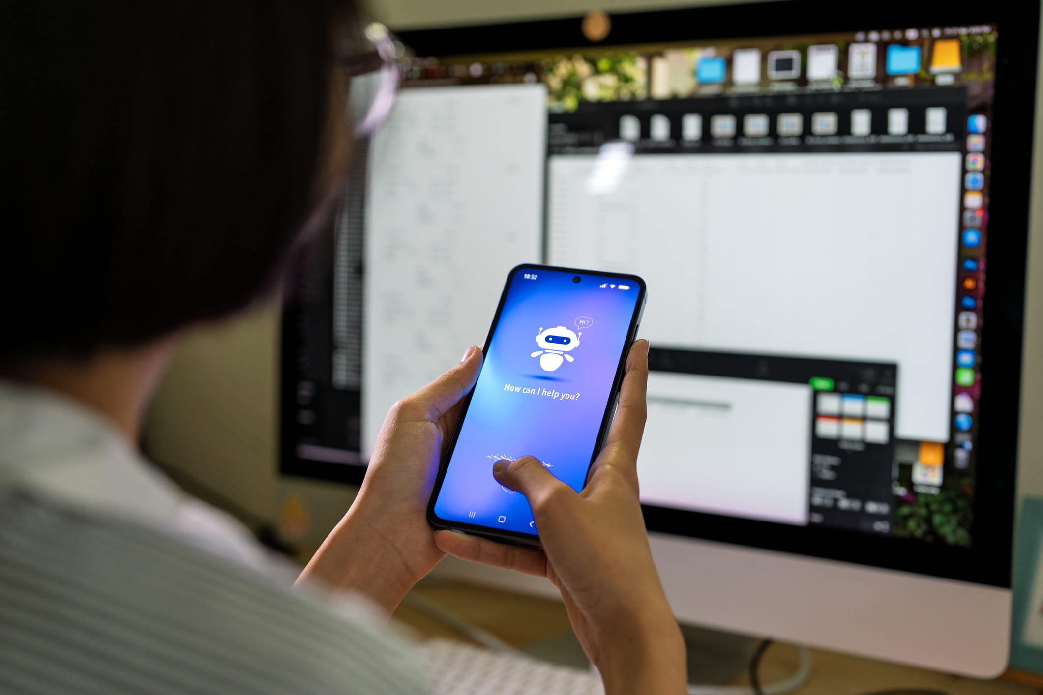 A person sitting in front of a computer, using an AI tool on their phone.