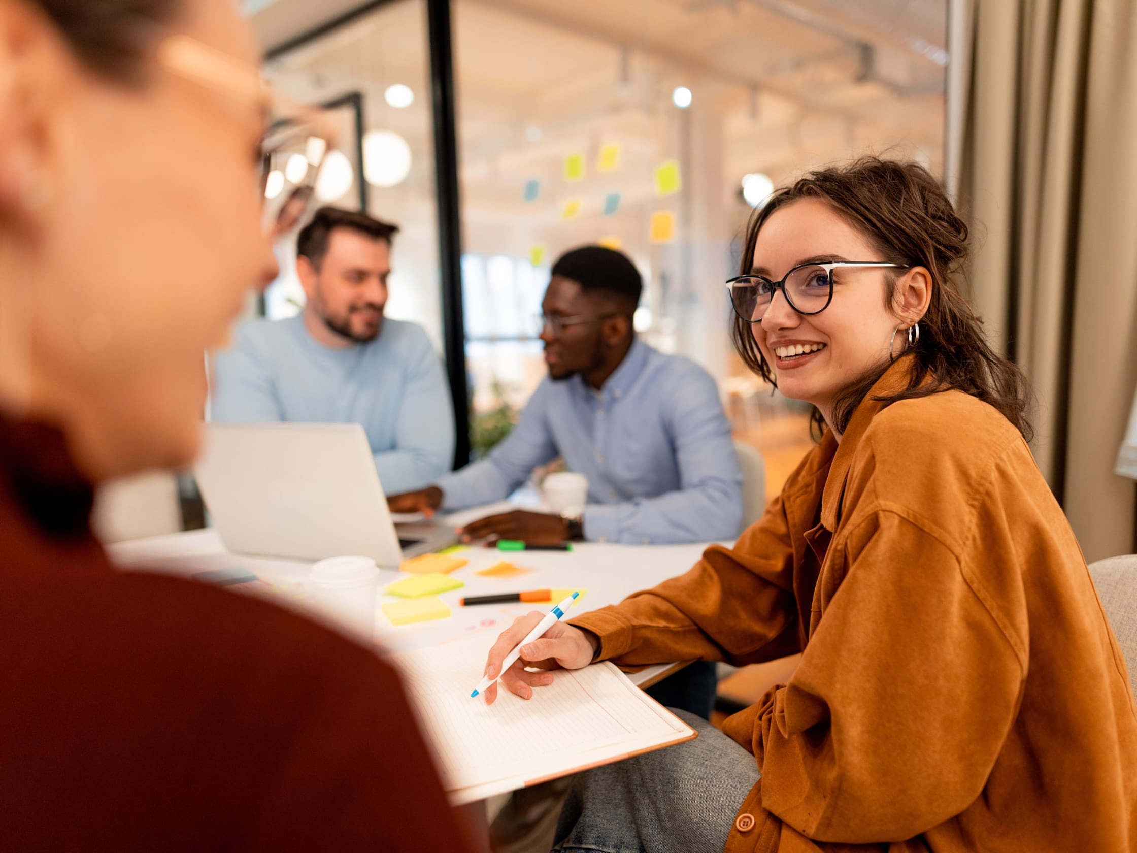 A group of people in an office, with notebooks and a laptop, smiling in conversation.
