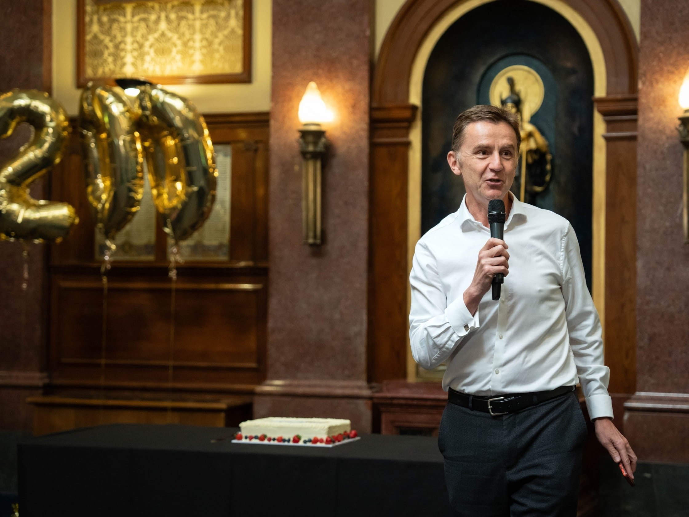 Ian Jeffery stands holding a microphone, in front of a cake and balloons in the shape of the number 200.
