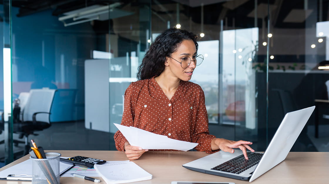 A woman with dark curly hair is sitting at a desk looking at a laptop. She is wearing a brown button up shirt and glasses. She is holding a piece of paper in her right hand. 