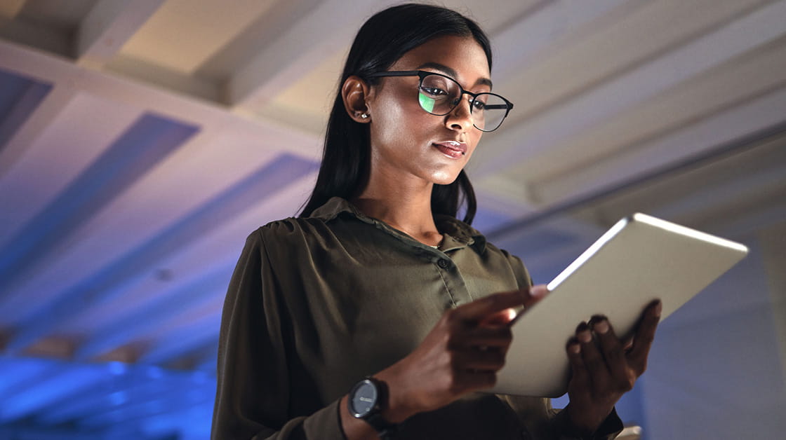 A woman with long dark hair and wearing glasses looks at a tablet that she is holding in her hands. The background is of a ceiling lit with purple lighting.