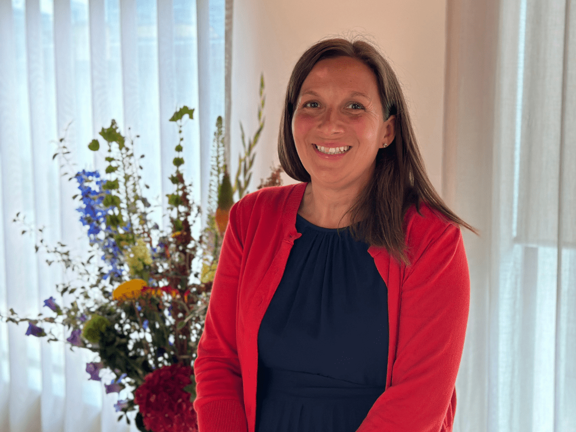 A smiling woman in a blue dress stands next to some flowers.