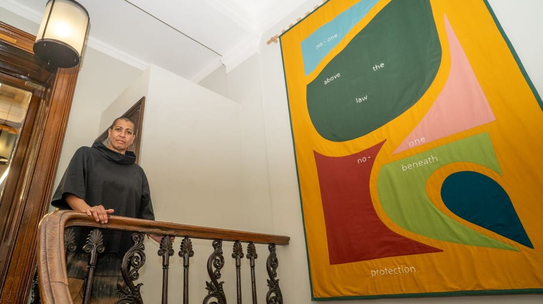 Helen Cammock stands at the top of a staircase next to a brightly coloured fabric banner hanging on a wall.