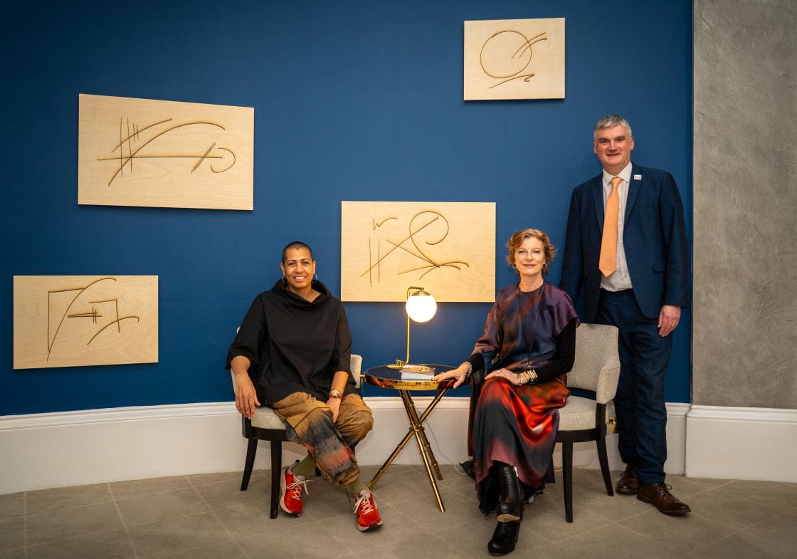 Helen Cammock (lef) and Fabienne Nicholas (middle) sit on chairs with Mark Evans standing to the right). They are positioned in front of four plywood art canvases.