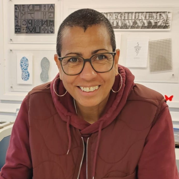 Artist Helen Cammock smiles while wearing a dark red hoodie. Helen is a black woman with short black hair and glasses