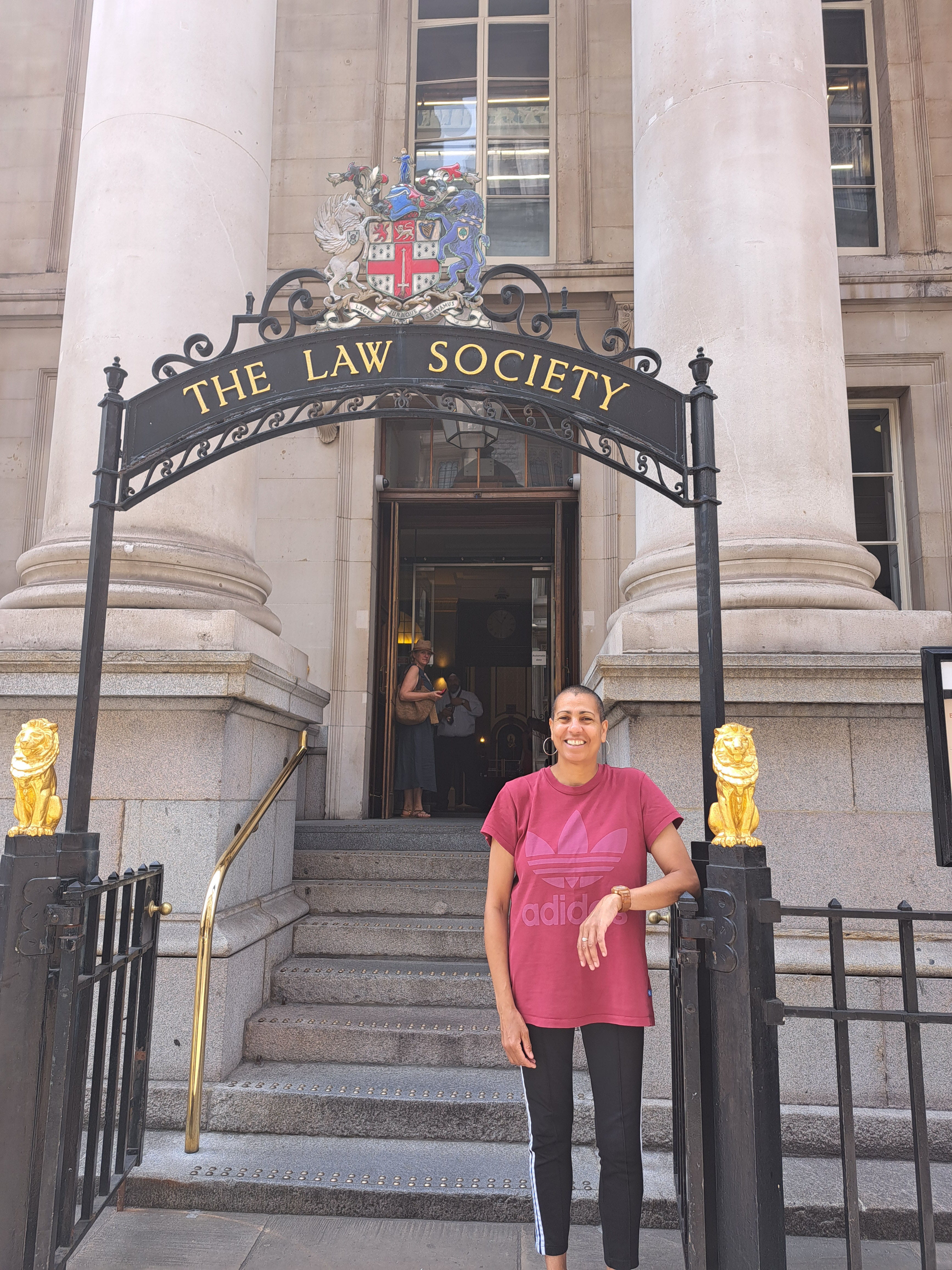 Artist Helen Cammock stands outside the Law Society building on Chancery Lane. Helen is a black woman with short black hair, she is smiling and wearing a dark red sports top with black leggings