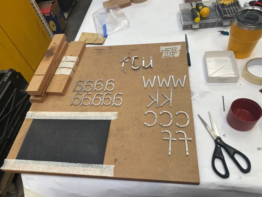Helen's art in progress a close up of a studio work bench that features several steel letters that will be used in the art on a wooden board. There's a piece of black sandpaper next to the letters and a set of scissors on the table.