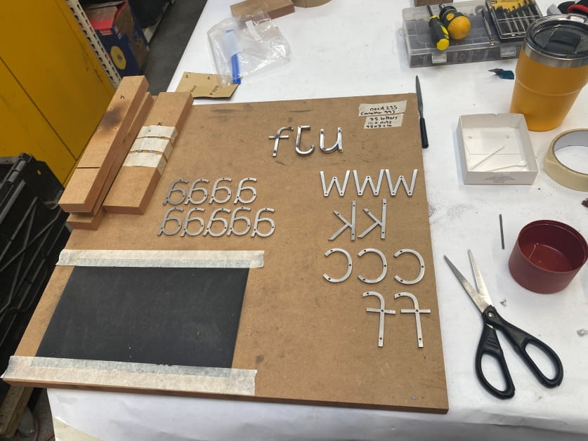 Helen's art in progress a close up of a studio work bench that features several steel letters that will be used in the art on a wooden board. There's a piece of black sandpaper next to the letters and a set of scissors on the table.
