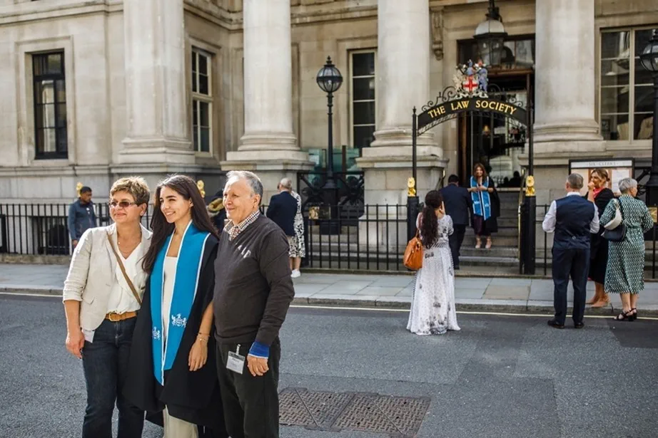 Two parents stand proudly with their daughter who is dressed in graduation robes outside the Law Society on an admission ceremony day. In the back of the photo more people are celebrating.