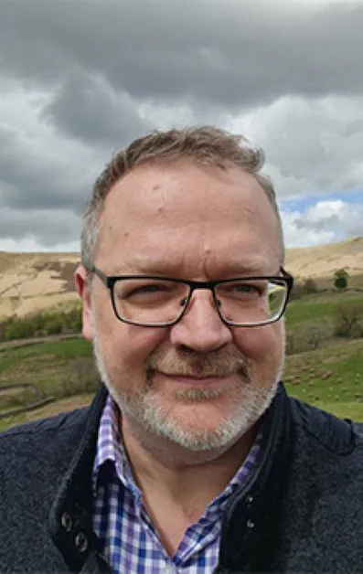 Person in glasses and a checkered shirt standing outdoors with scenic hills in the background.