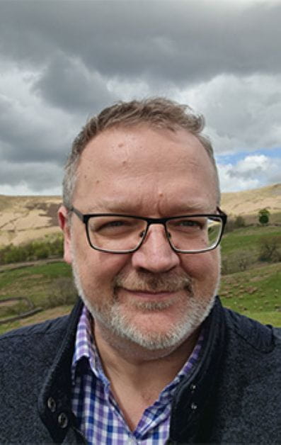 Person in glasses and a checkered shirt standing outdoors with scenic hills in the background.