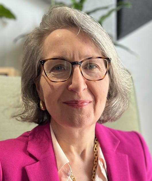 Person with short hair and glasses wearing a pink jacket, smiling at the camera.