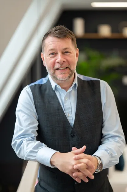 Person stands with hands clasped, wearing a light blue shirt and dark grey waistcoat.