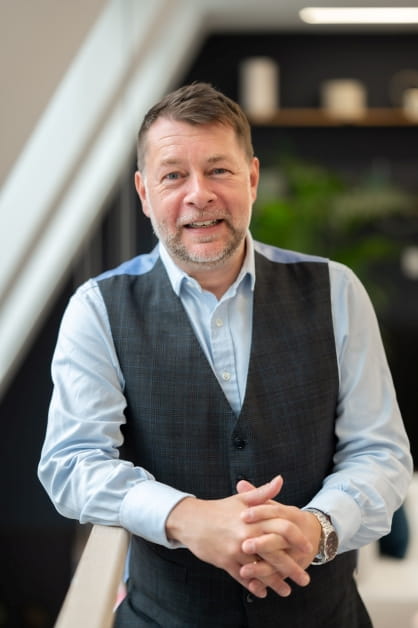 Person stands with hands clasped, wearing a light blue shirt and dark grey waistcoat.