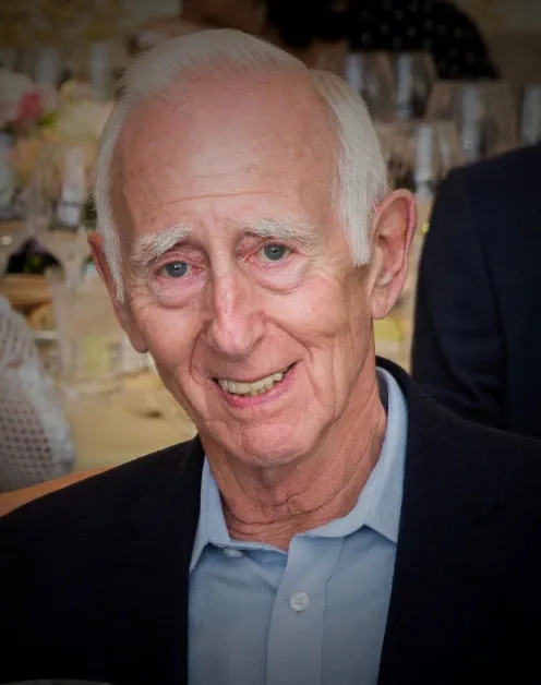 Older individual with short white hair smiling indoors in a casual setting.