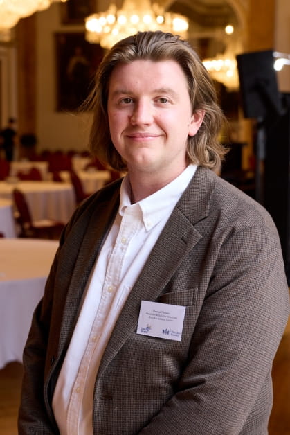 Junior solicitor smiles at an event whilst wearing a white shirt and smart brown jacket. 