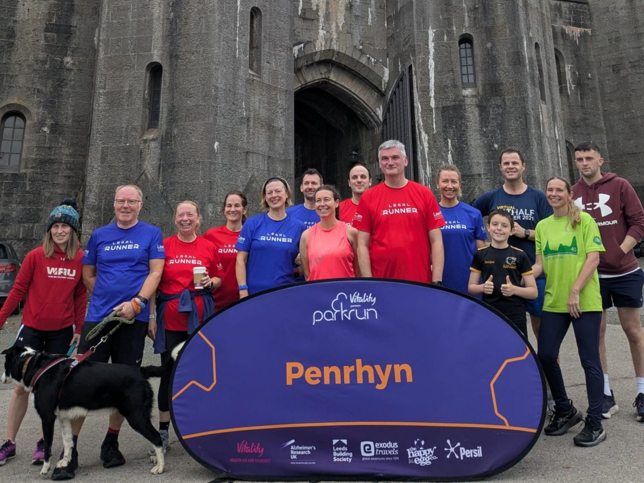 Mark stands with fellow parkrunners outside Penrhyn Castle in North Wales. They've placed a large sign in front of them that reads 'Penrhyn'.