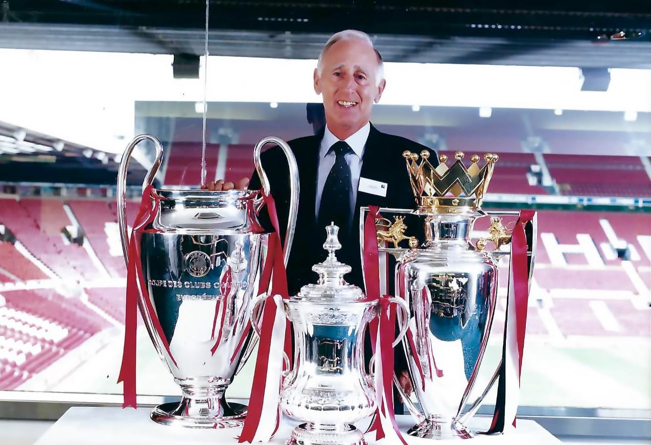 A white man in a suit stands smiling in a stadium next to three large football trophies