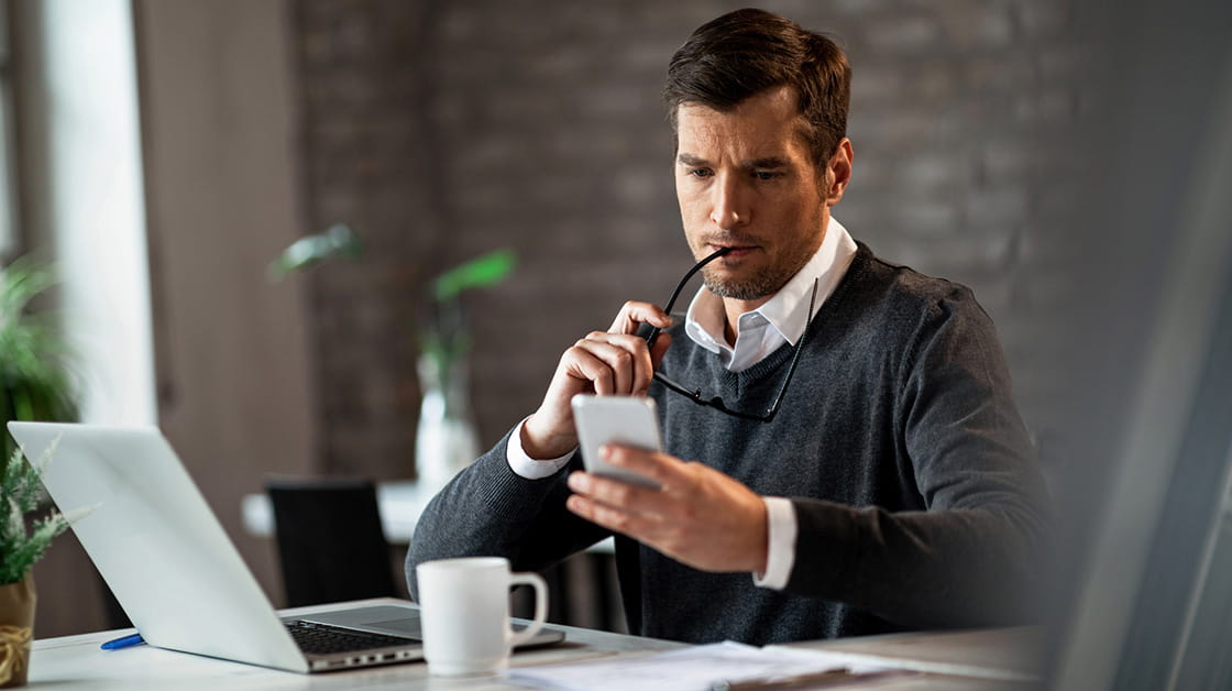 A man in business dress sits at an office desk and reads from his mobile phone.