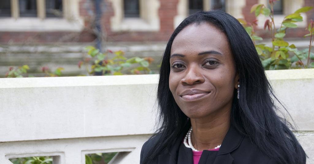 Saida Bello is a black woman with shoulder length straight hair. Behind her is an old red-brick building and rose bushes