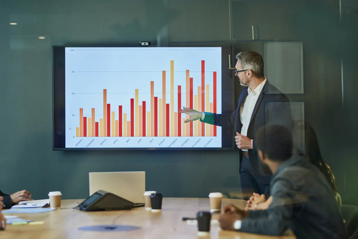 Businessman presenting financial statistics to diverse group of colleagues during meeting in modern boardroom. Professionals discussing data and graphs on a large screen.