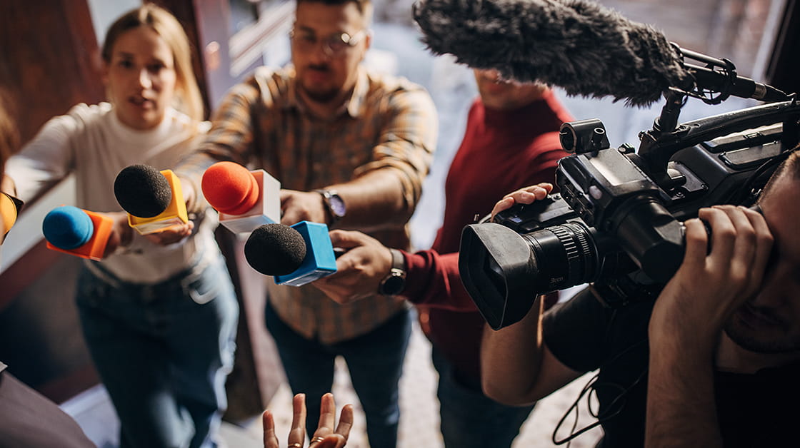 A group of journalists stands on a doorstep, holding microphones and cameras up to ask questions.
