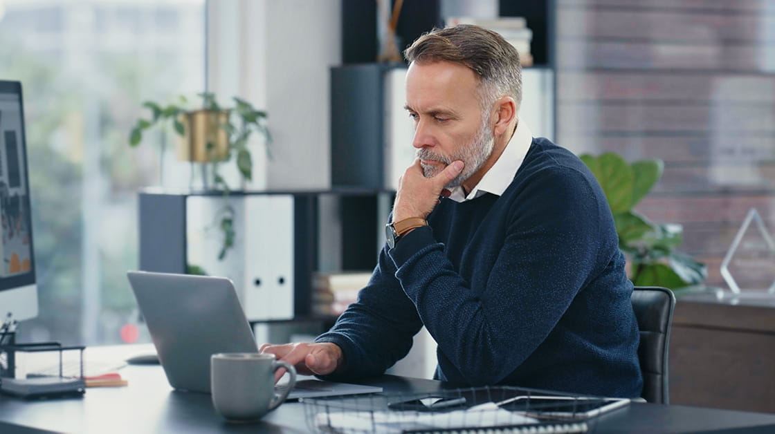 A white man with grey hair and a beard sits at a desk considering his finances. He wears a white shirt and navy jumper.
